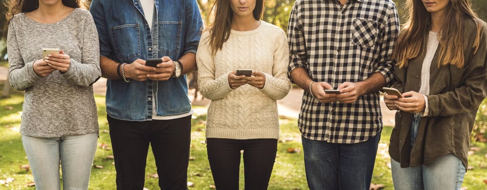 Five people standing outdoors, all looking down and using their smartphones; only their torsos and hands are visible, highlighting brand engagement in today’s connected world.